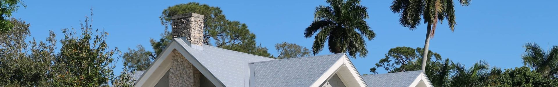 Zweistöckiges graues Einfamilienhaus mit hellem Satteldach, Veranda und Kamin, umgeben von Rasen, Sträuchern und Palmen